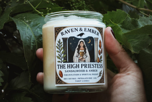 Hand holding a candle labeled 'The High Priestess' by Raven & Ember against a leafy background