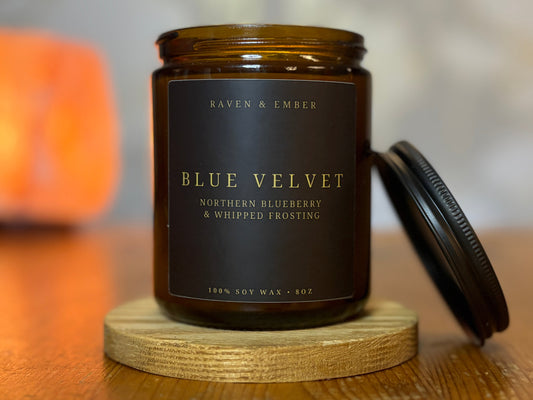 Candle jar labeled 'Blue Velvet' by Raven & Ember on a wooden surface with a blurred background.
