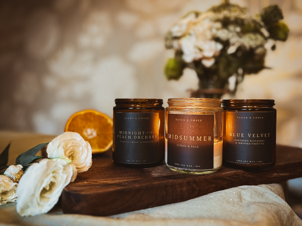 Three scented candles in amber glass jars arranged on a wooden board with white flowers and an orange slice. The candles are labeled Midnight Peach Orchard, Midsummer, and Blue Velvet.