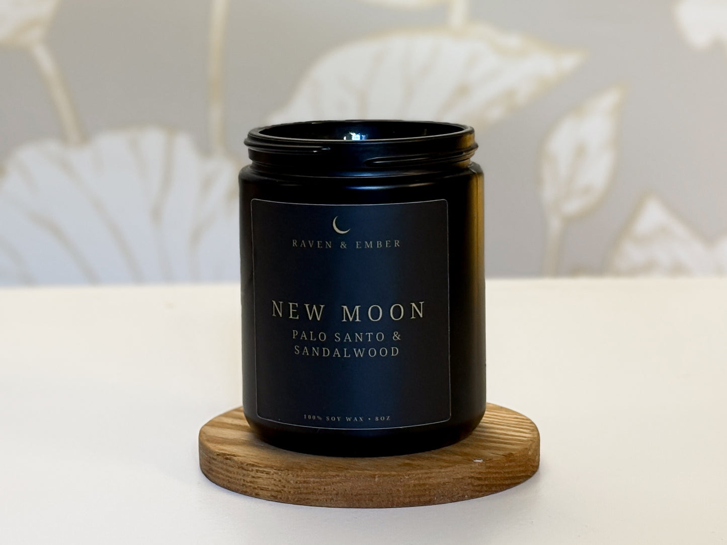 Black candle jar labeled 'New Moon' with Palo Santo & Sandalwood on a wooden coaster.