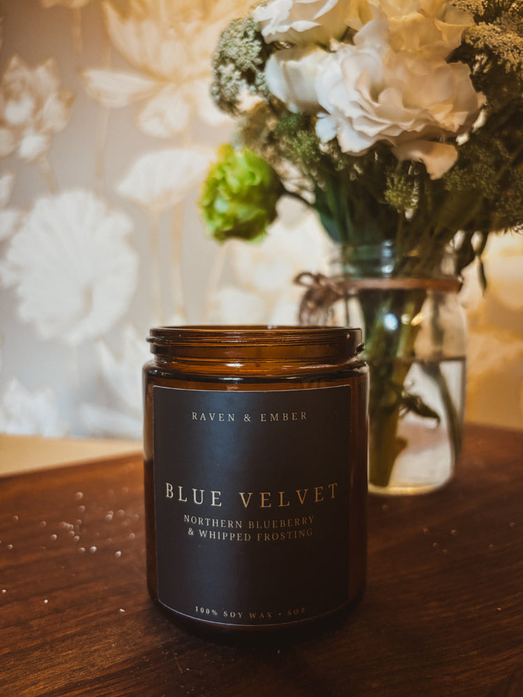 An amber glass jar candle with a dark brown label reading 'BLUE VELVET' by Raven & Ember, styled on a wooden surface with a blurred floral arrangement in the background.